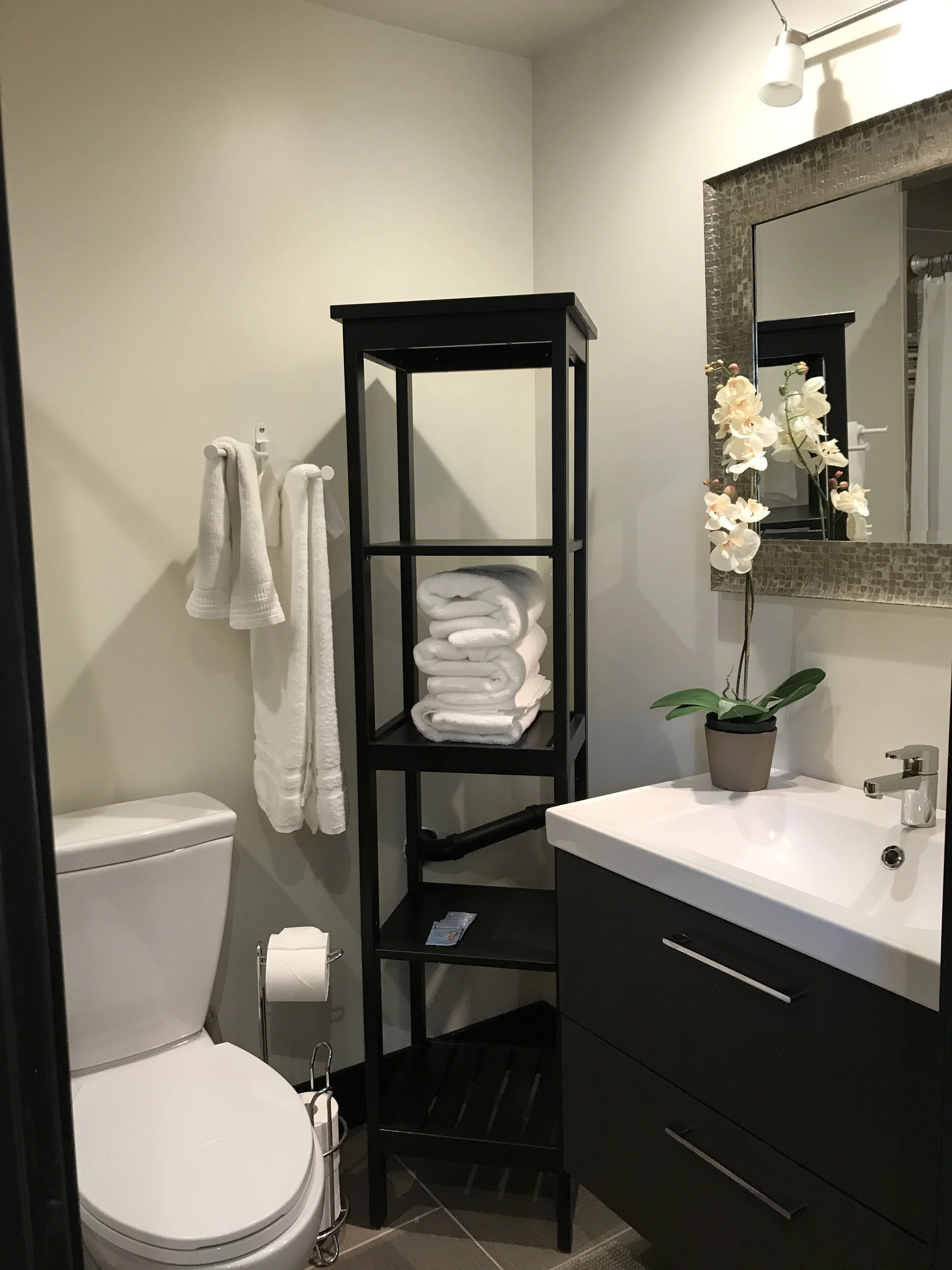 A bathroom with a toilet, a black shelving unit holding white towels, a sink with a black vanity, a large mirror, and a potted orchid.
