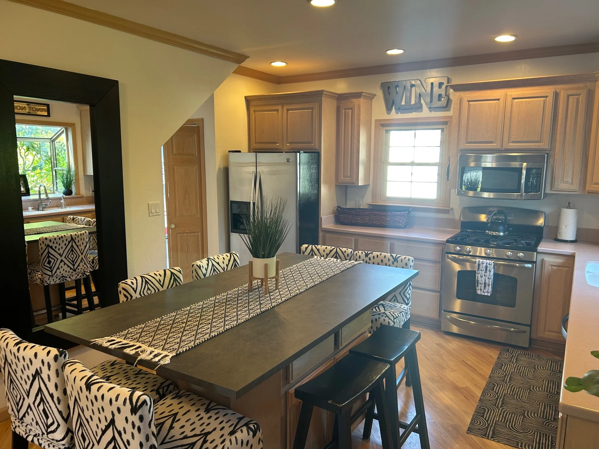 A modern kitchen with wooden cabinets, stainless steel appliances, a large black dining table with patterned chairs, a decorative plant, and a window above the sink, illuminated by ceiling lights.