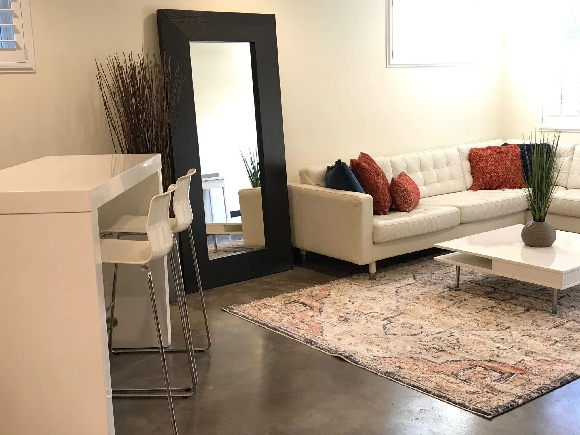 Living room with white sofa, red and black cushions, white coffee table with a vase of tall grass, a large black-framed mirror, and a bar-height white counter with two white chairs.