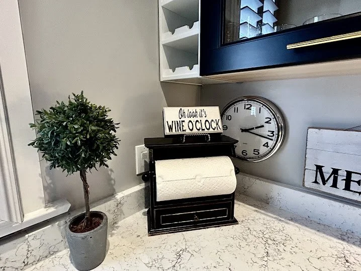 Kitchen countertop with a potted plant, a paper towel holder, a sign reading "Oh look it's WINE O CLOCK," a wall clock, and a part of a wooden sign with the letters "ME".