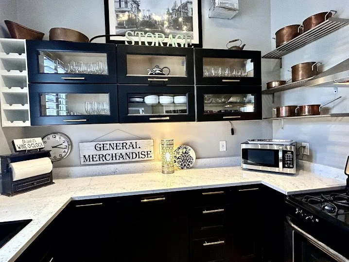 Kitchen countertop with black cabinets, open shelves with copper pots, microwave, and decorative items including a clock, sign reading 'GENERAL MERCHANDISE', and a glass jar with a candle. There's a window with a street view behind the upper cabinets.