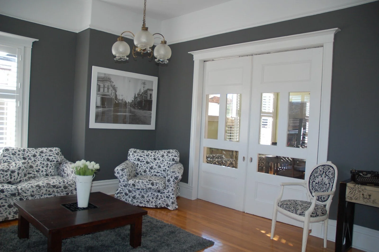 Living room with dark gray walls, white trim, wooden floor, white patterned sofa and armchair, wooden coffee table with a white vase of flowers, framed black and white photograph on the wall, glass-paneled white sliding door, and a vintage chair with a floral cushion.