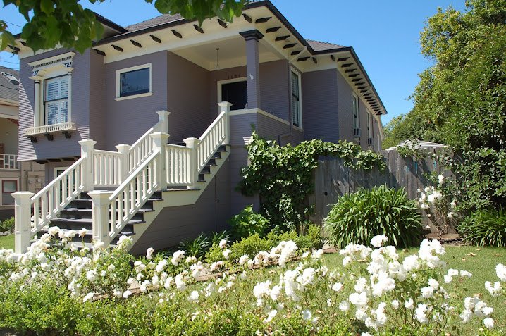 A two-story house with a staircase leading up to a front porch, surrounded by a lush green garden with white flowers and bushes, under a clear blue sky.