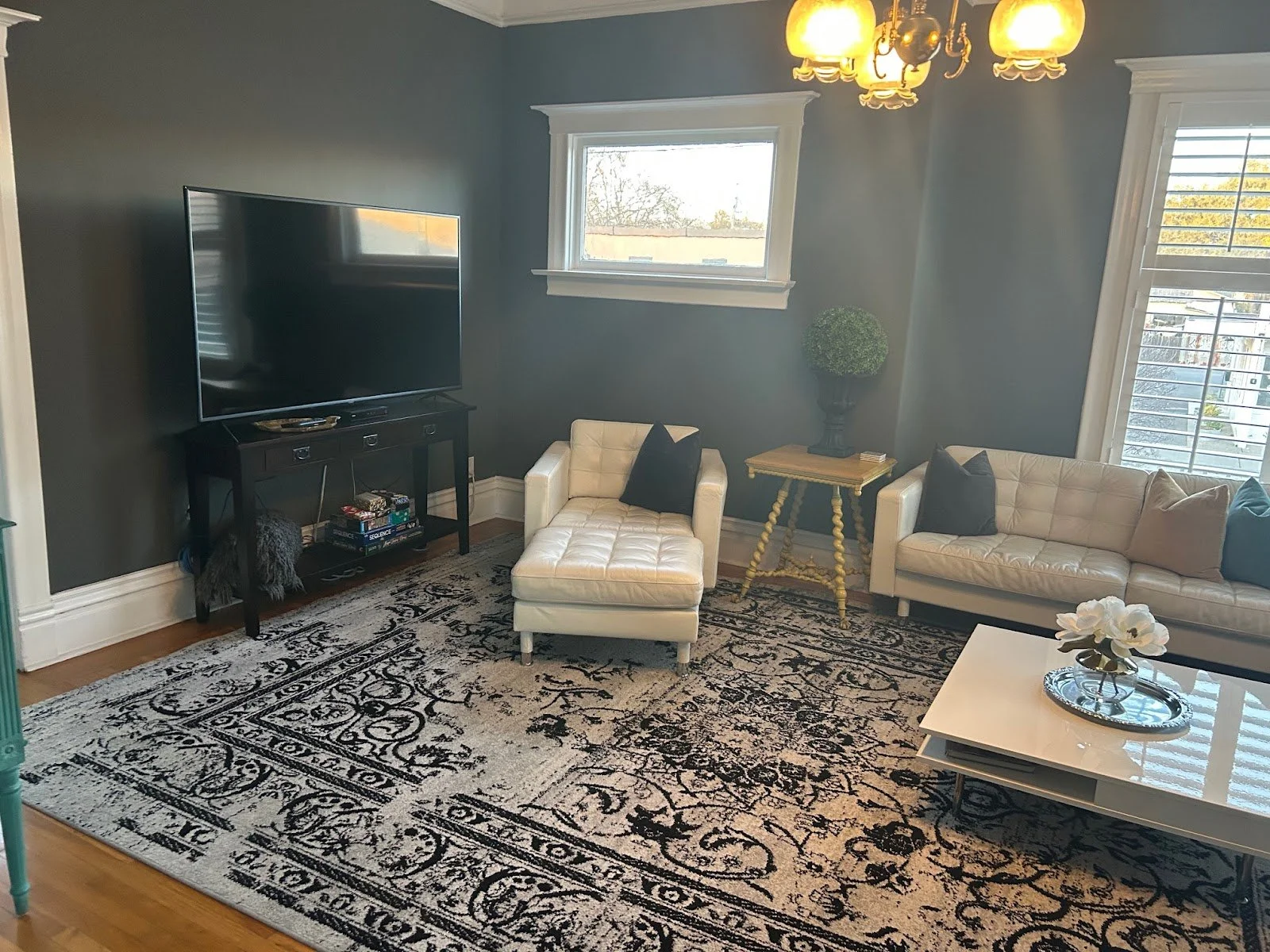 Living room with a large patterned black and white rug, a white armchair with a black pillow, a beige sofa with black and beige pillows, a black TV on a black stand, a gold side table with a potted plant, a white coffee table with a vase of white flowers, a window with shutters, and a gold chandelier overhead.