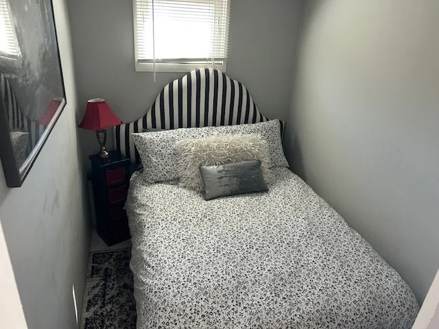 A small bedroom with a bed against the wall, a colorful headboard, a window with blinds, a nightstand with a pink lampshade, and patterned bedding with decorative pillows.
