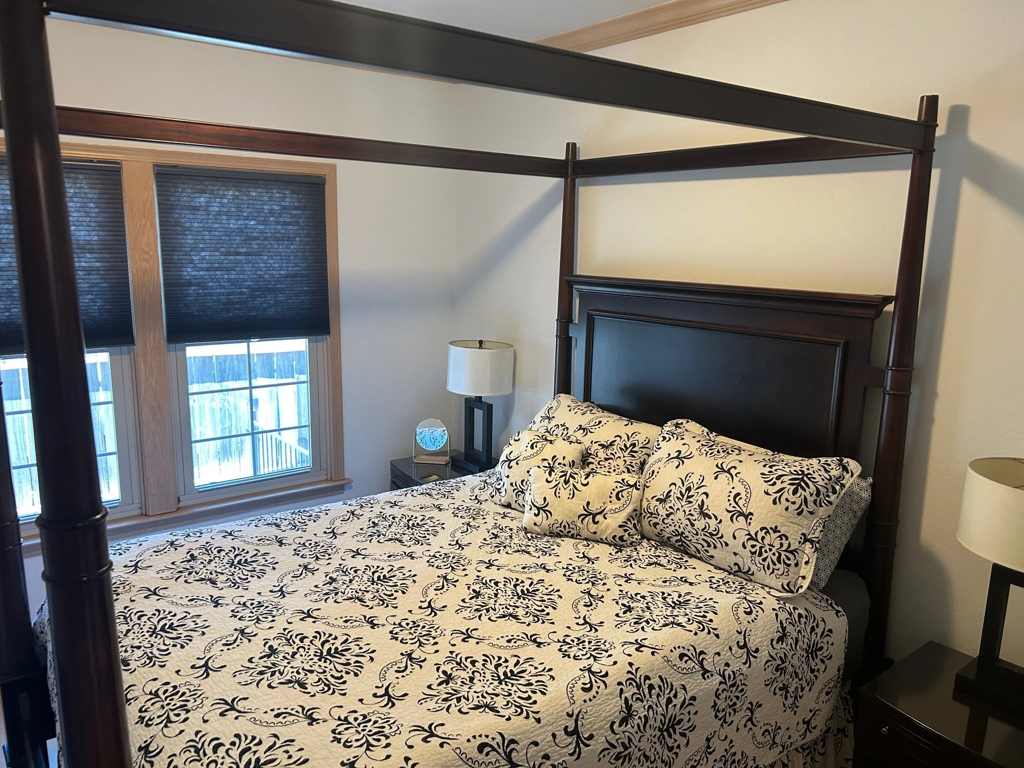 Bedroom with a dark wooden canopy bed, a patterned bedspread, two pillows, and two nightstands with lamps. Two windows with dark curtains are above one nightstand.