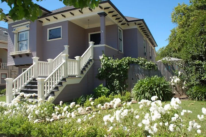 A two-story Victorian house with a staircase leading to the front porch, surrounded by a garden with white flowers and green foliage, under a blue sky.