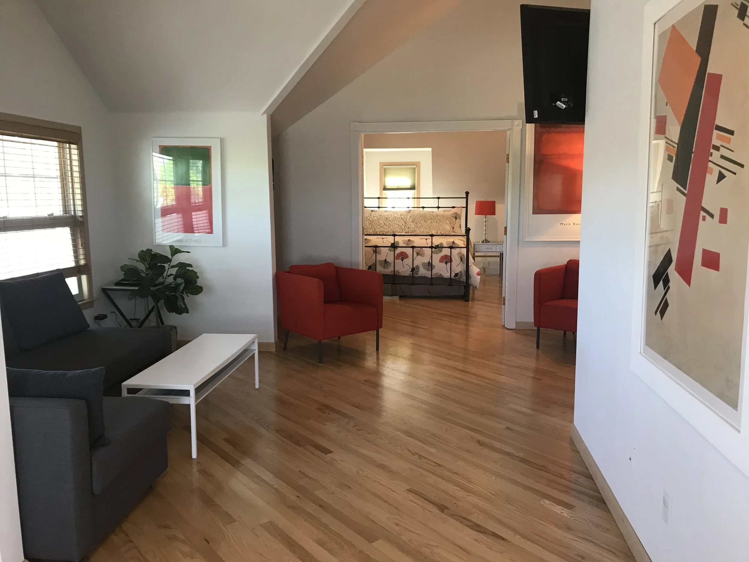 Living room with hardwood floors, gray and red chairs, large windows with blinds, and colorful artwork on the walls. A glimpse into a bedroom with a metal bed frame and a bedside table with a red lamp.