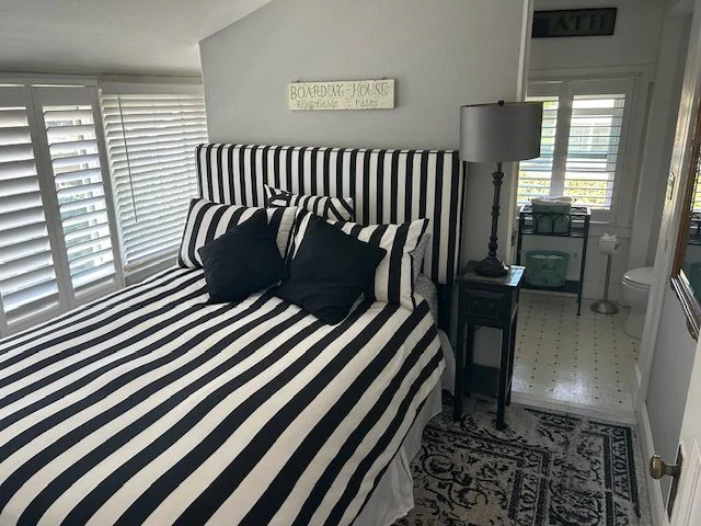 A bedroom with black and white striped bedding, pillows, and headboard. There is a window with blinds, a bedside table with a lamp, and a view of a small bathroom with a toilet and window.