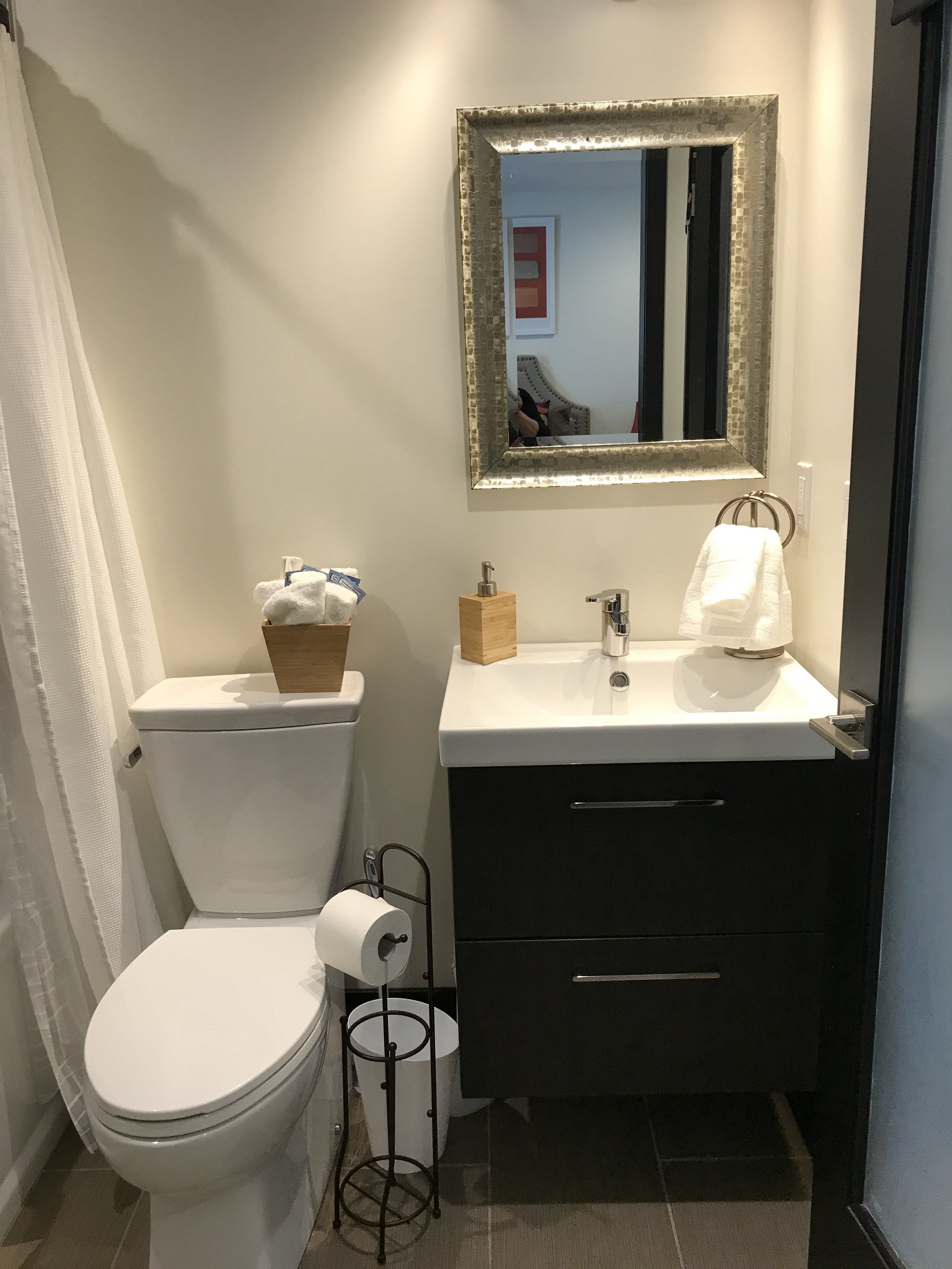 A small bathroom with a toilet, a dark vanity with a sink, a mirror, and a small wooden tray with towels on the toilet tank. There is a toilet paper holder and a soap dispenser on the vanity, and a towel hanging on a ring near the mirror.