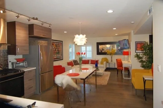 Open-concept living and dining area with wood flooring, modern furniture, and colorful decor, including a white sofa with red pillows, a yellow armchair, a dining table with red plates and tulips, and a kitchen corner with stainless steel appliances.
