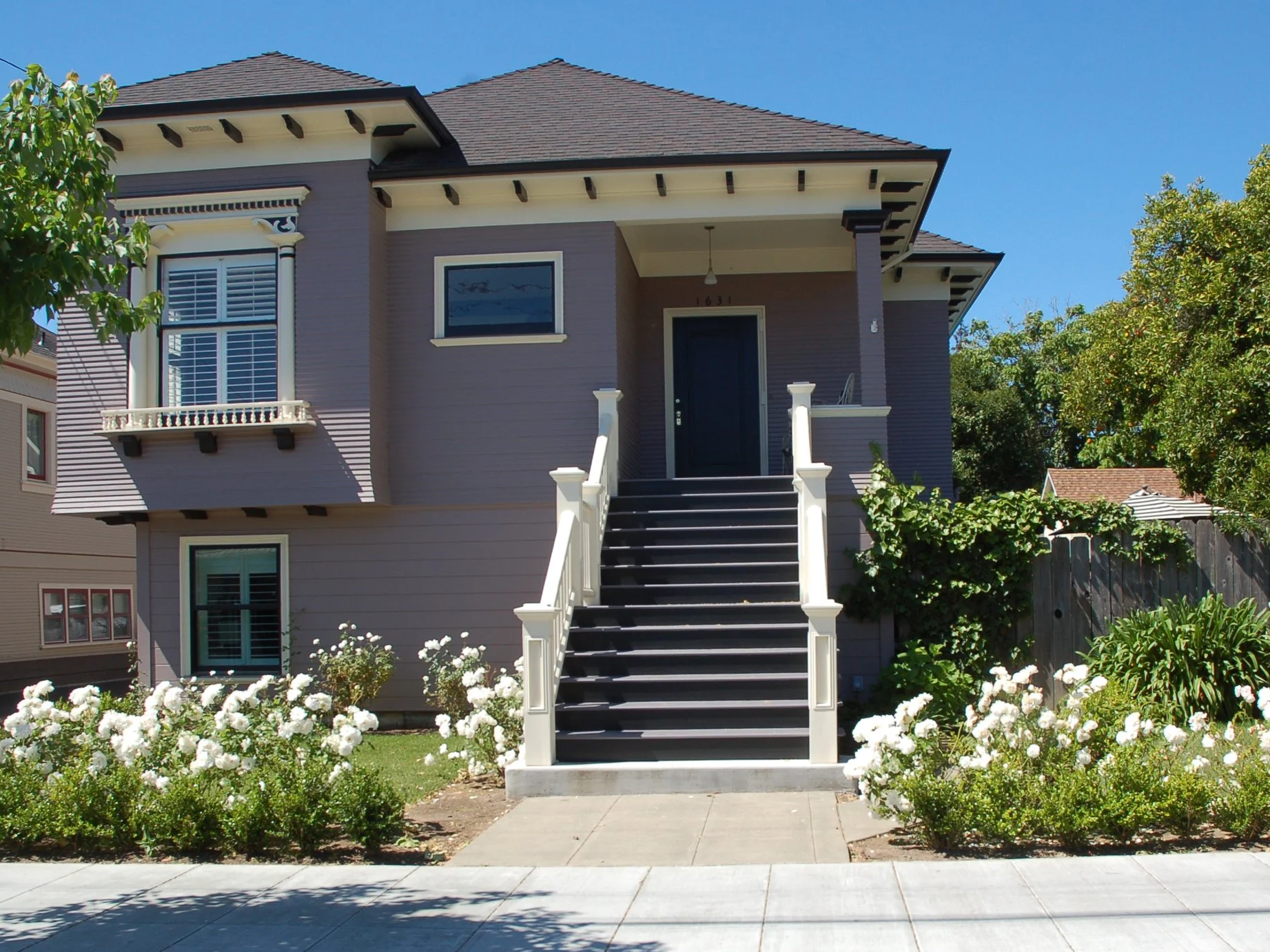 A two-story house painted in purple with white trim, front staircase, and a small porch, surrounded by garden with white flowers and green bushes.