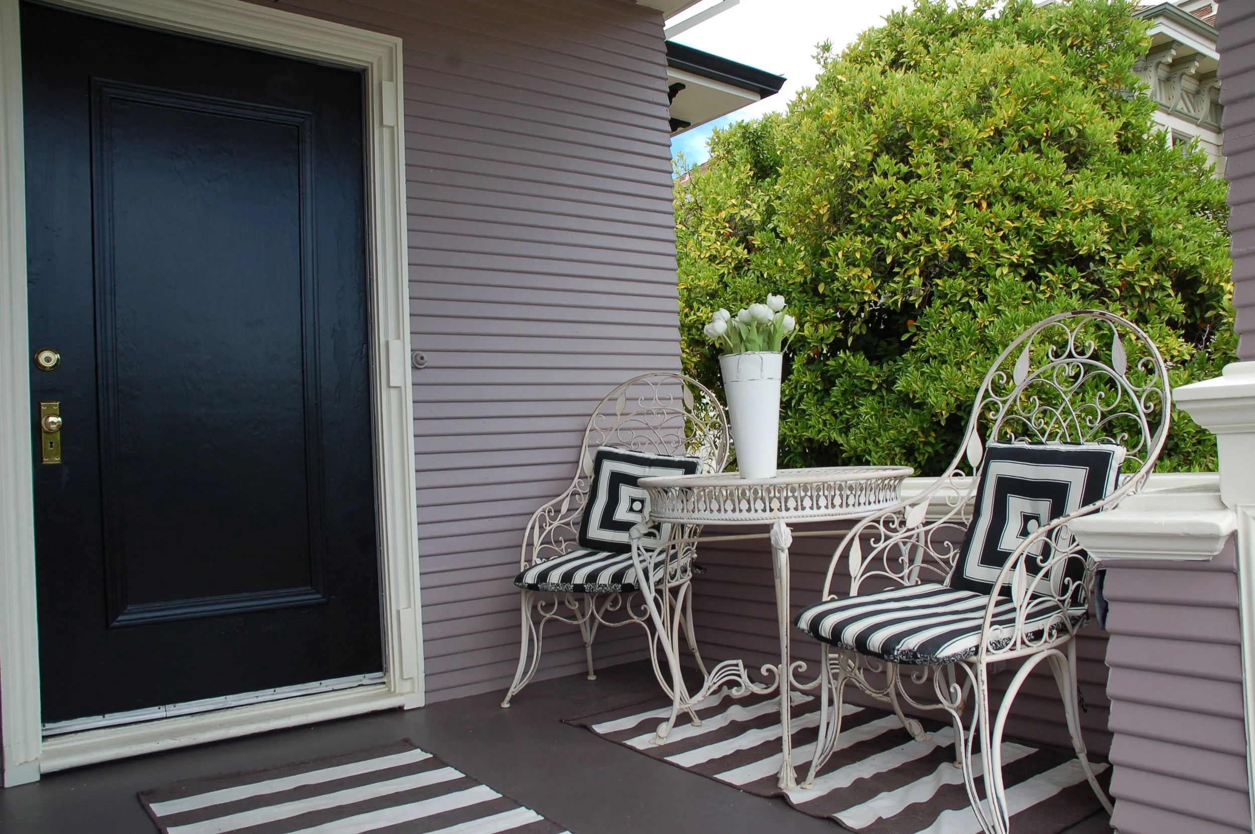 A porch with a black door, striped rugs, white wrought iron chairs with black and white cushions, a white table with a vase of white tulips, and green bushes in the background.