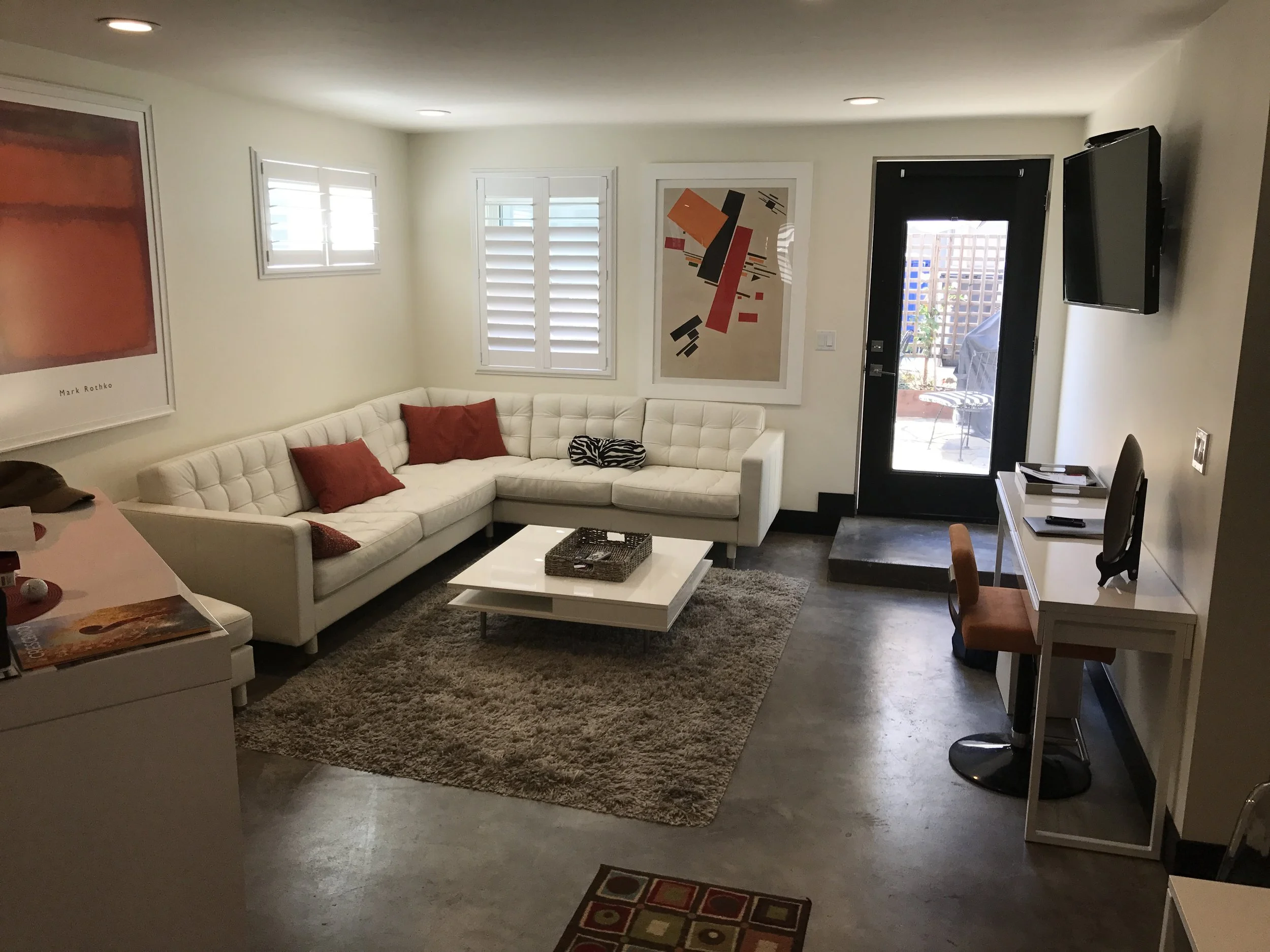 Living room with white sectional sofa, red and black cushions, abstract artwork on the walls, a coffee table on a beige rug, a mounted TV, a white desk with a brown chair, and glass door leading outside to a patio.