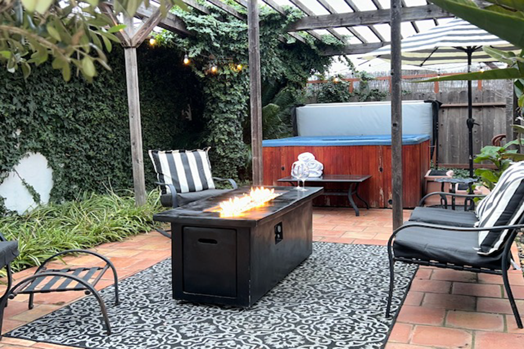 Outdoor patio with a fire table, lounge chairs with striped cushions, a hot tub, umbrella, and string lights, surrounded by lush greenery and a wooden fence.