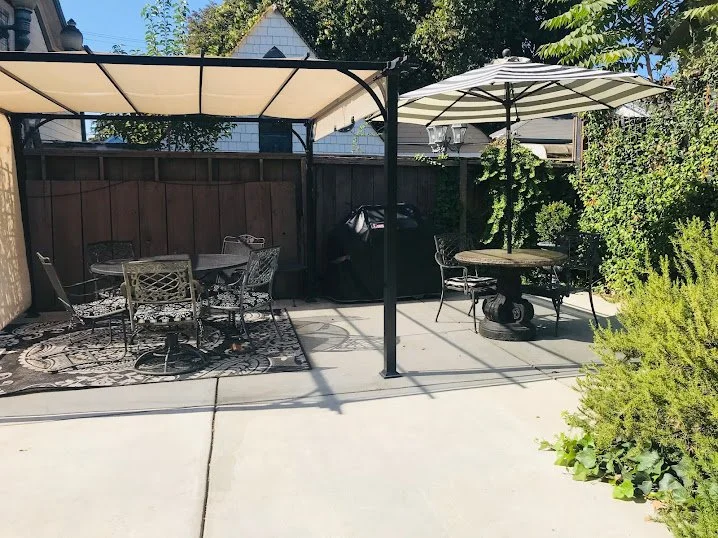 Backyard patio area with two patio tables and chairs, umbrellas for shade, a grill, and greenery around the perimeter.