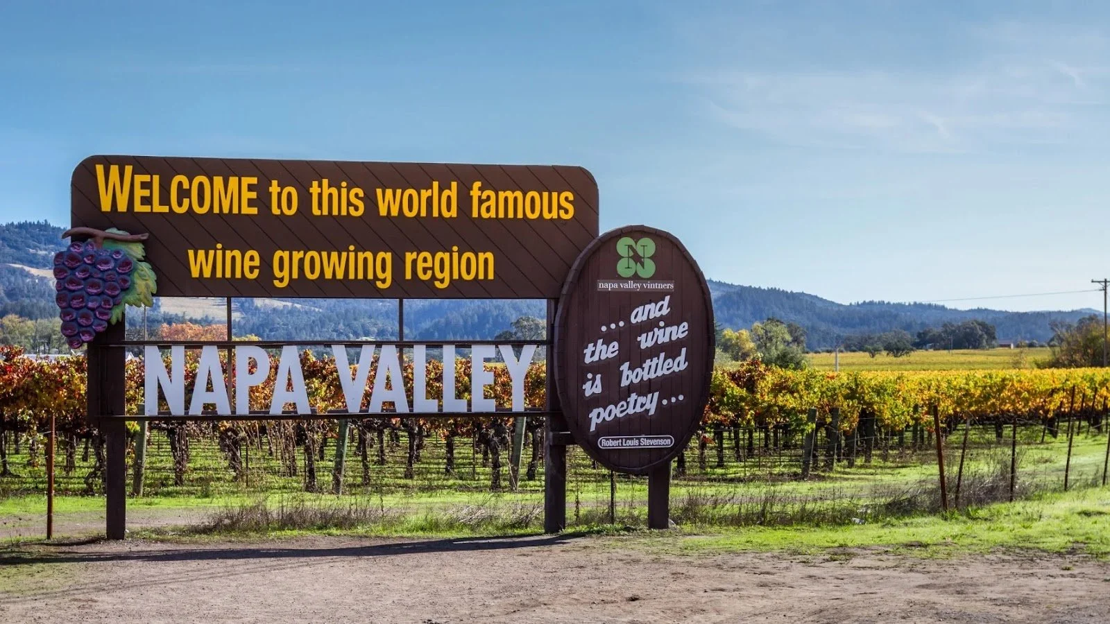 A large sign welcomes visitors to Napa Valley, a famous wine region. The sign features a bunch of grapes on the left and includes a quote about wine. The background shows rows of vineyards with hills and a clear blue sky.
