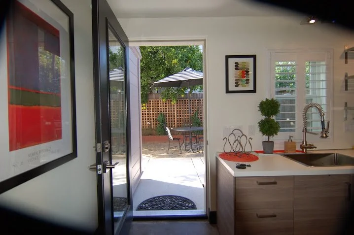 View from inside a kitchen looking out to a backyard patio with a table, chairs, and umbrella, through an open door.