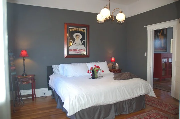 A bedroom with dark gray walls and hardwood floors. A large bed with white bedding and pillows, and a brown fur throw. Two small nightstands with red lamps on each side of the bed. Framed vintage posters on the walls. A chandelier hanging from the ceiling.