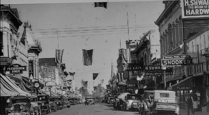 Black and white photo of a busy downtown street with vintage cars and storefronts, including a shoe store, flower shop, and an ad for a snooker hall.