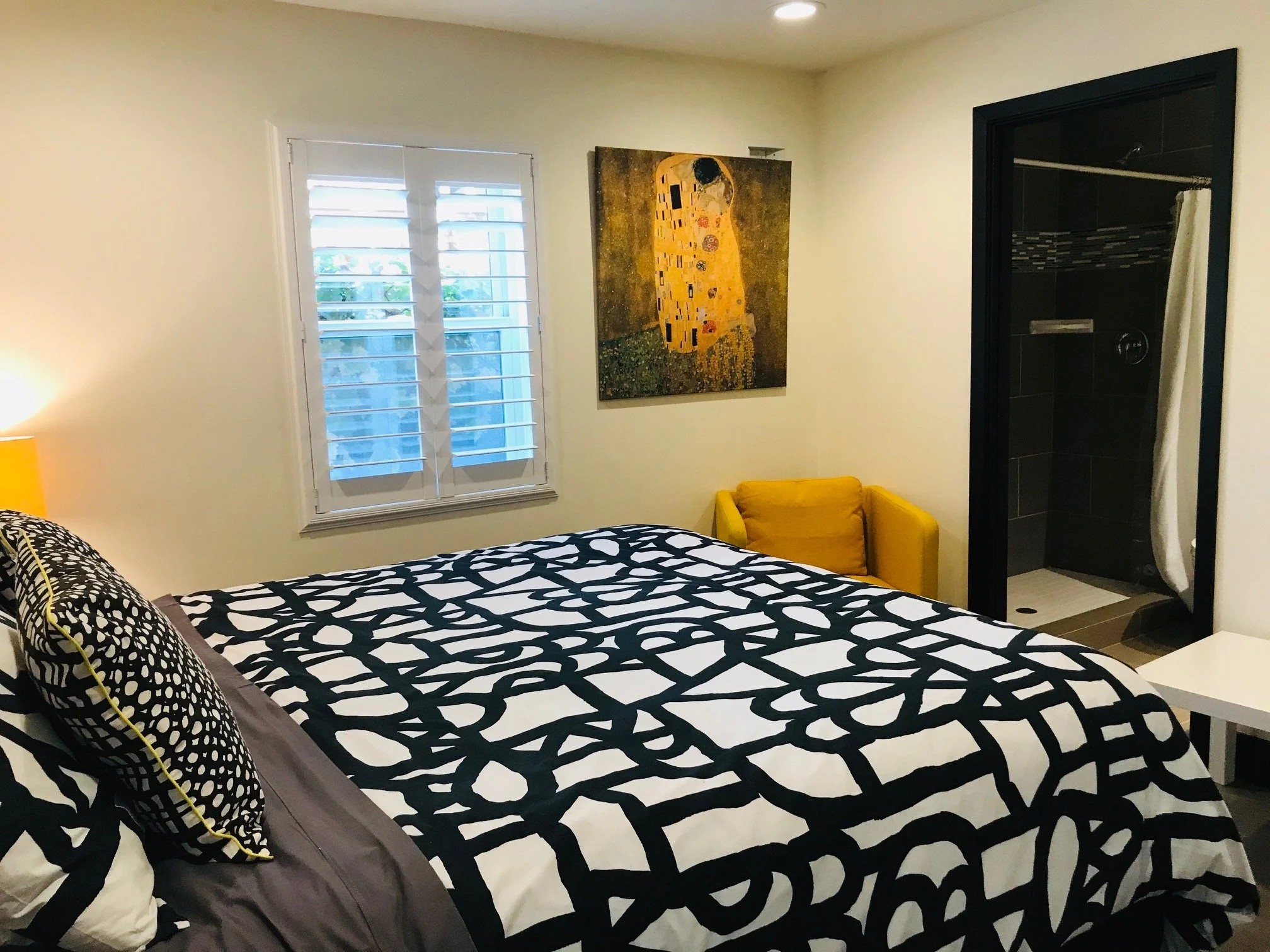 A bedroom with a bed that has a black and white geometric patterned bedding and pillows, a yellow armchair, a window with white blinds, a colorful abstract painting, and an open shower area with black tiles.