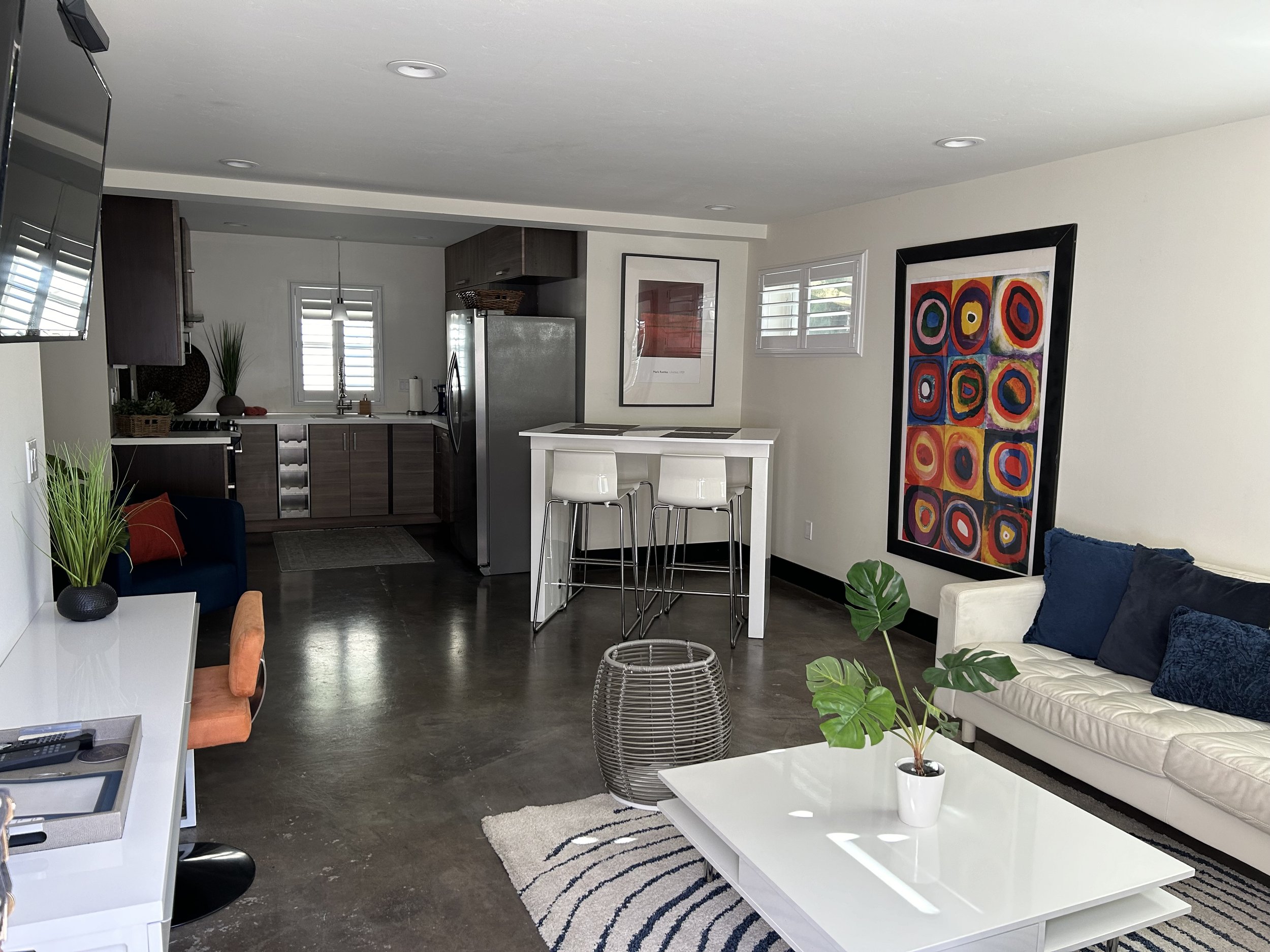 Living room with white couch and blue cushions, white coffee table with potted plant, colorful abstract wall art, dark hardwood floors, modern kitchen with stainless steel appliances, high chairs at a breakfast bar, and decorative items and plants around the space.