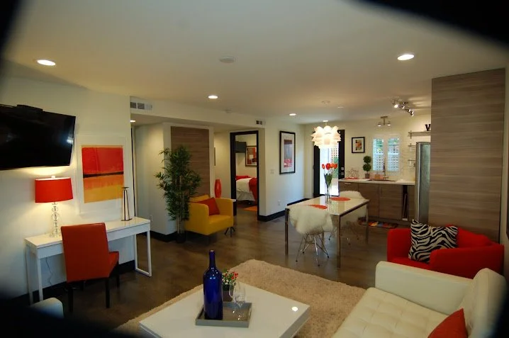 Open-concept living room and kitchen with modern furniture, a flat-screen TV, red, yellow, and white seating, decorative lamps, and indoor plants.
