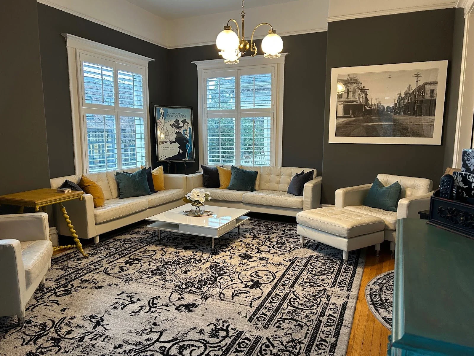 A living room with white sofas, a white armchair with an ottoman, and a black and white patterned rug. There are yellow, navy, and green cushions on the sofas. A white coffee table with a flower arrangement is in the center. The room has three large windows with white shutters, a chandelier with three globe lights, a black and white wall art, and a large framed black and white photograph on a dark gray wall.