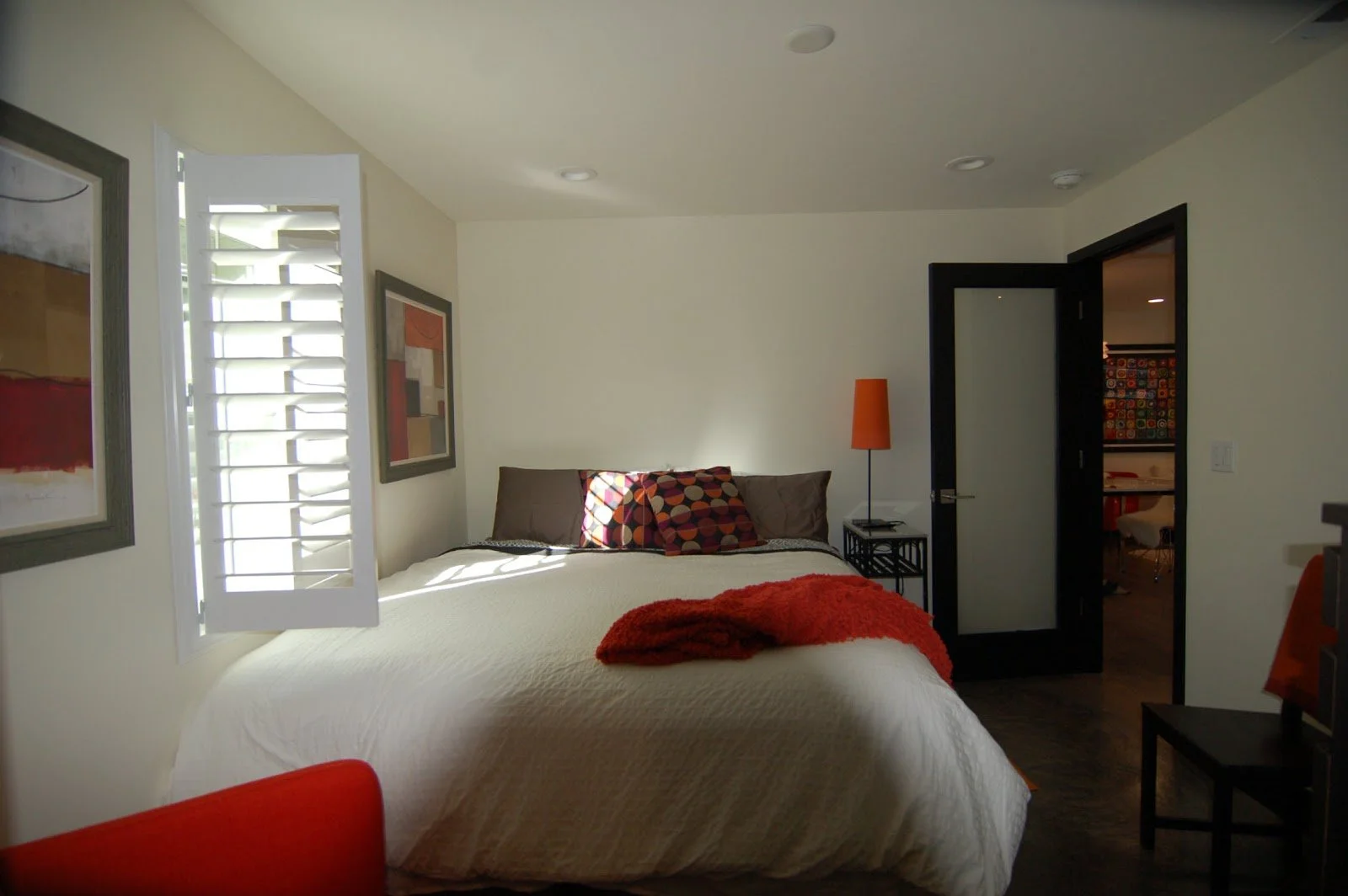 Bedroom with a bed, pillows, orange blanket, open window with shutters, artwork on the wall, black door, and a small table with an orange lamp.