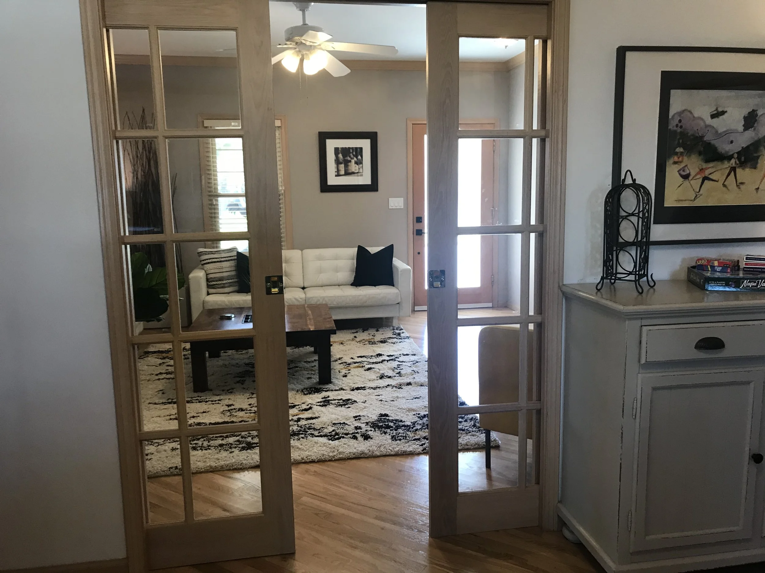 Living room seen through wooden-framed glass French doors, with a white sofa, a black pillow, a wooden coffee table on a patterned rug, and a windowed front door in the background.