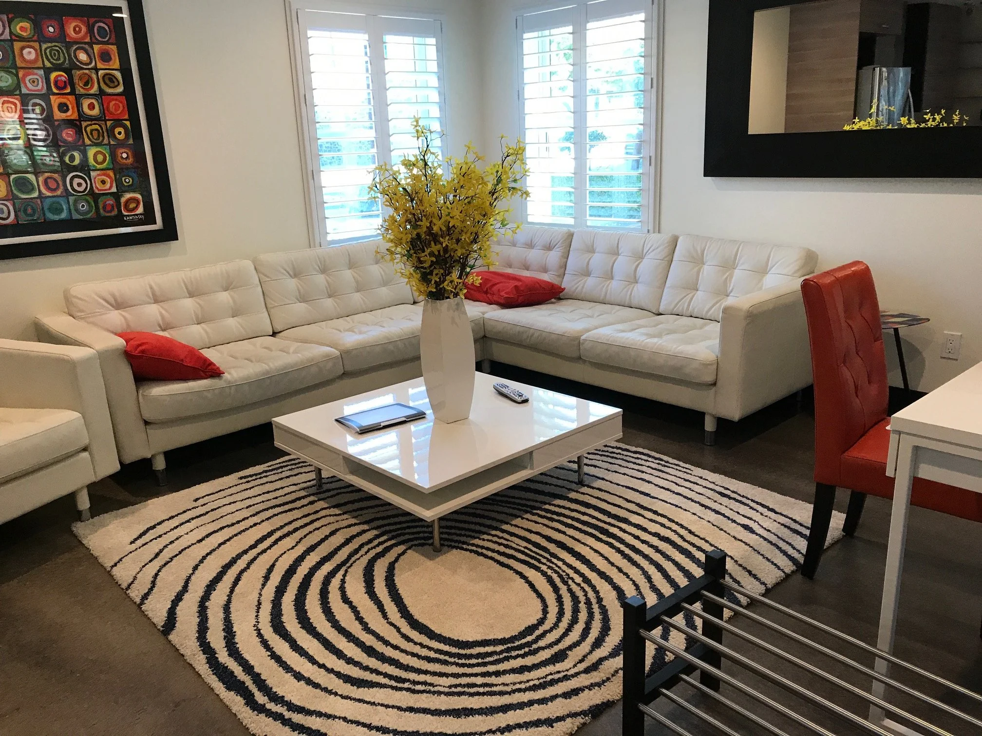 Living room with a white L-shaped sofa, red pillows, a white coffee table with a tablet and remote, a yellow floral centerpiece, a patterned rug, artwork on the wall, and window shutters.