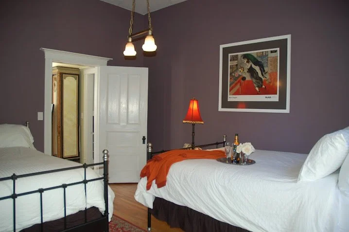 A bedroom with purple walls, two beds with white bedding, a bedside lamp with a red shade, a framed abstract art piece on the wall, and an open door leading to another room or closet.