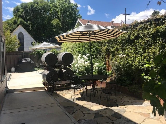 Outdoor patio with striped umbrellas, a table with four chairs, and whiskey barrels, surrounded by lush greenery and a small church building in the background.