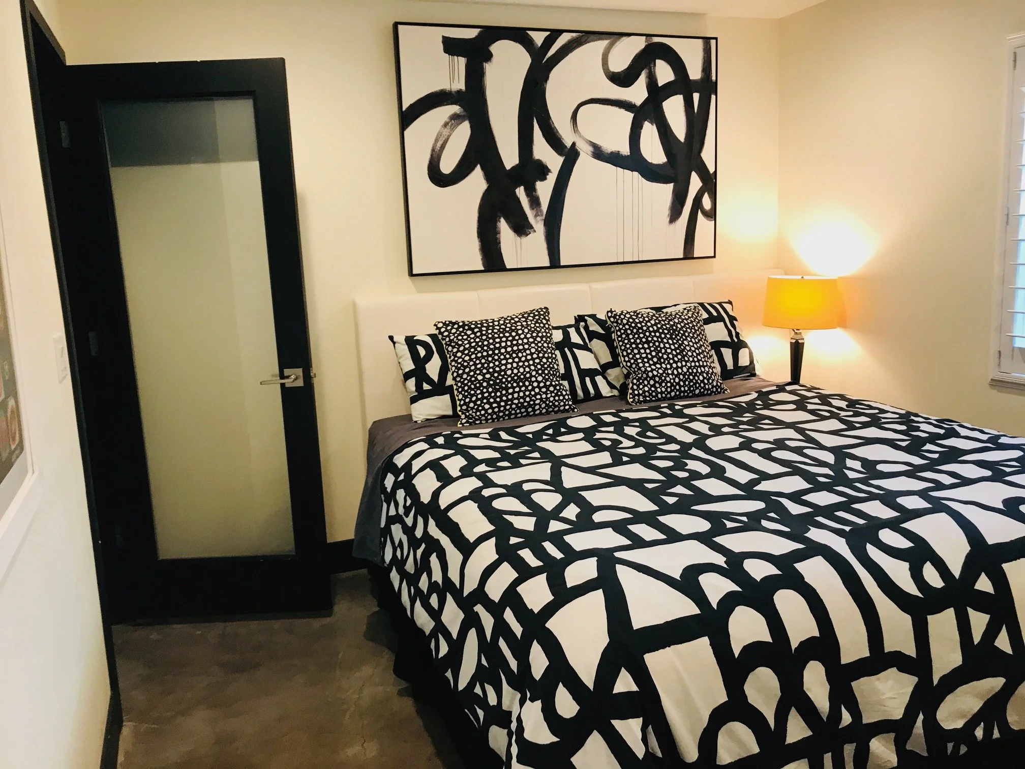 A bedroom with a black and white abstract theme. It features a bed with a black and white patterned duvet and pillows, a nightstand with a yellow lamp, a large abstract painting above the bed, and a window with white blinds.