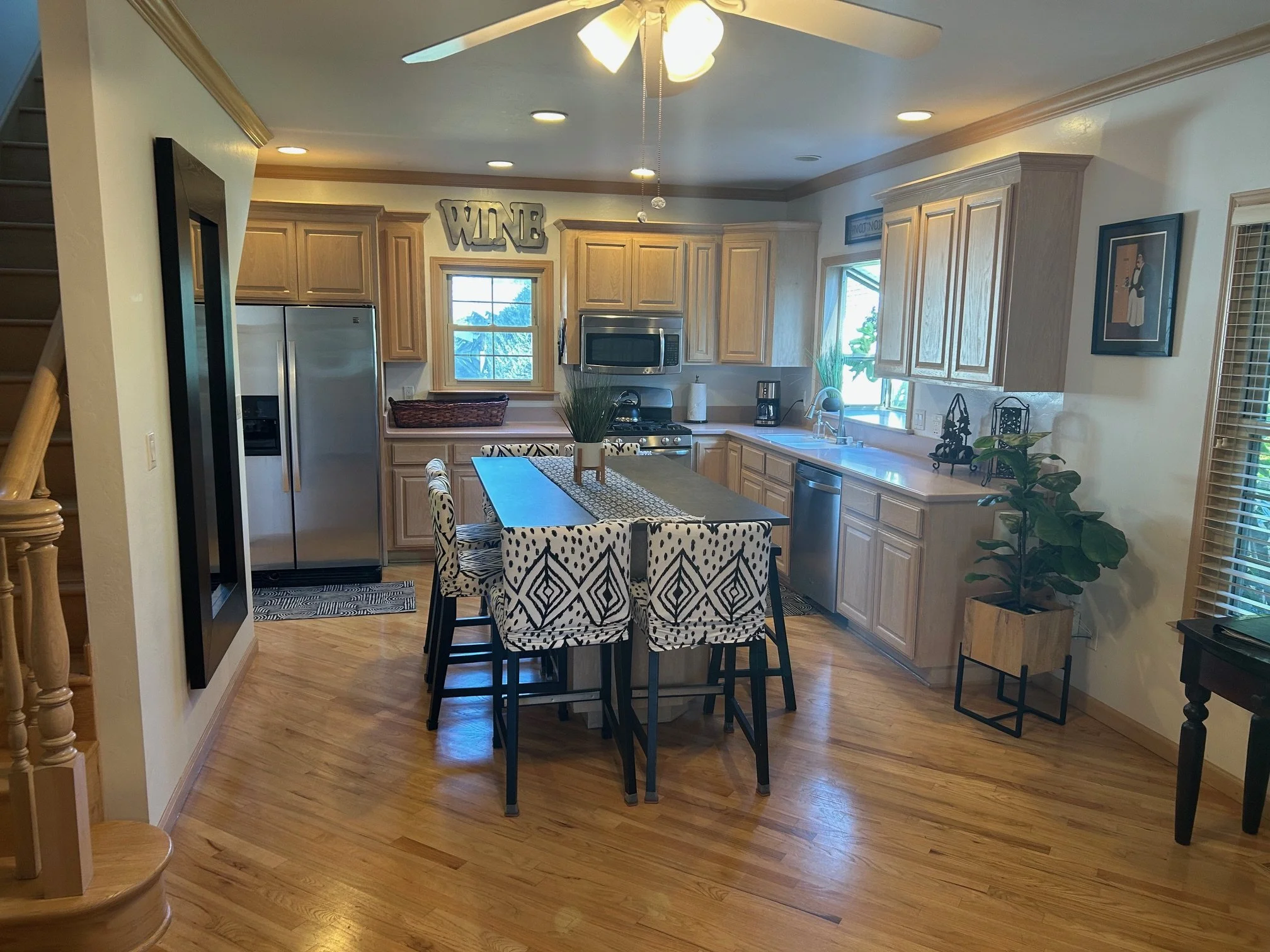 Bright kitchen with wooden cabinets, stainless steel refrigerator, microwave, and dishwasher. Small window above the sink, dining table with patterned chairs, and decorative items including a plant, a wine sign, and framed art.