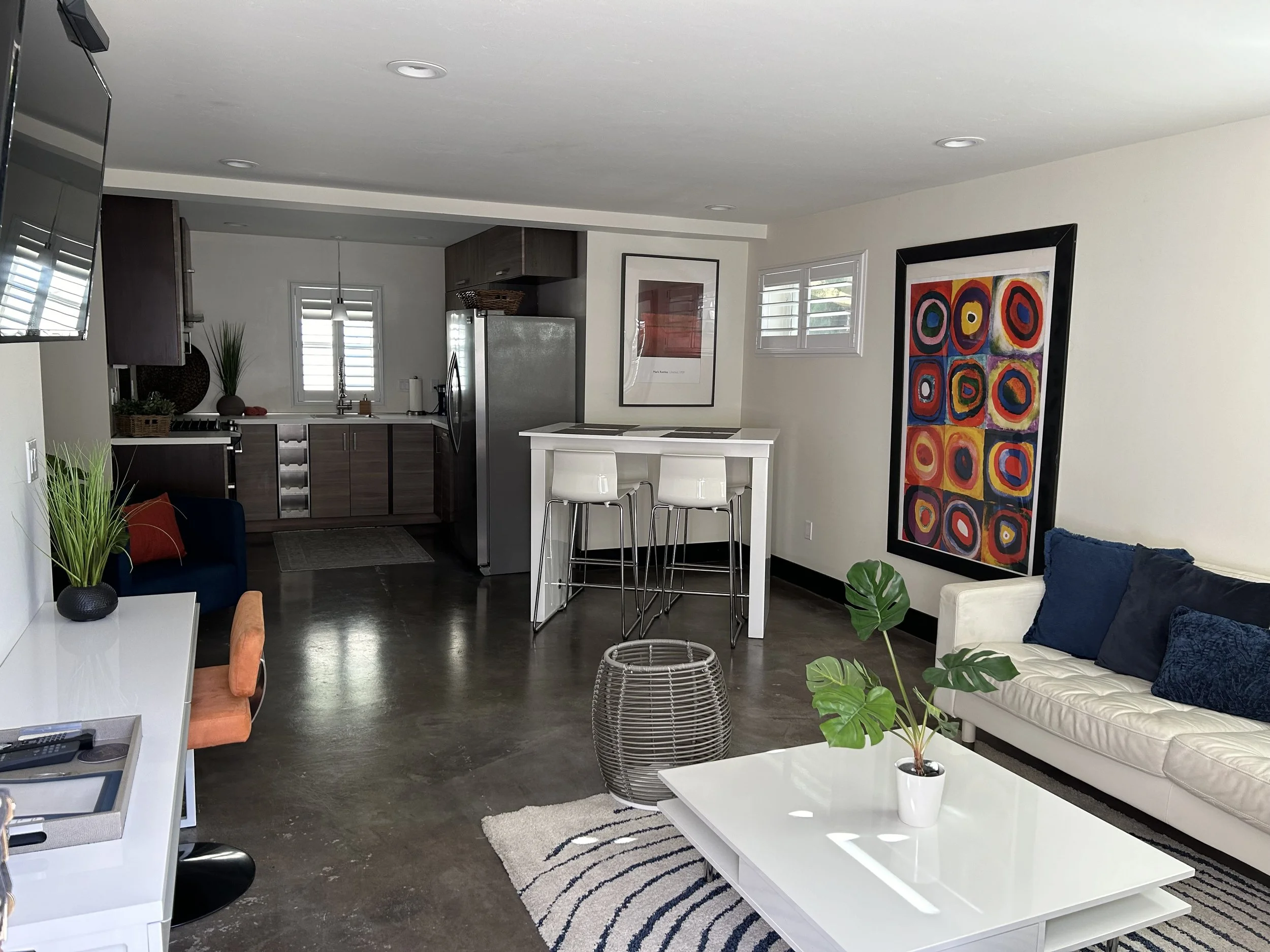 Open-concept living room and kitchen with modern decor, white walls, dark wood floors, and colorful artwork.