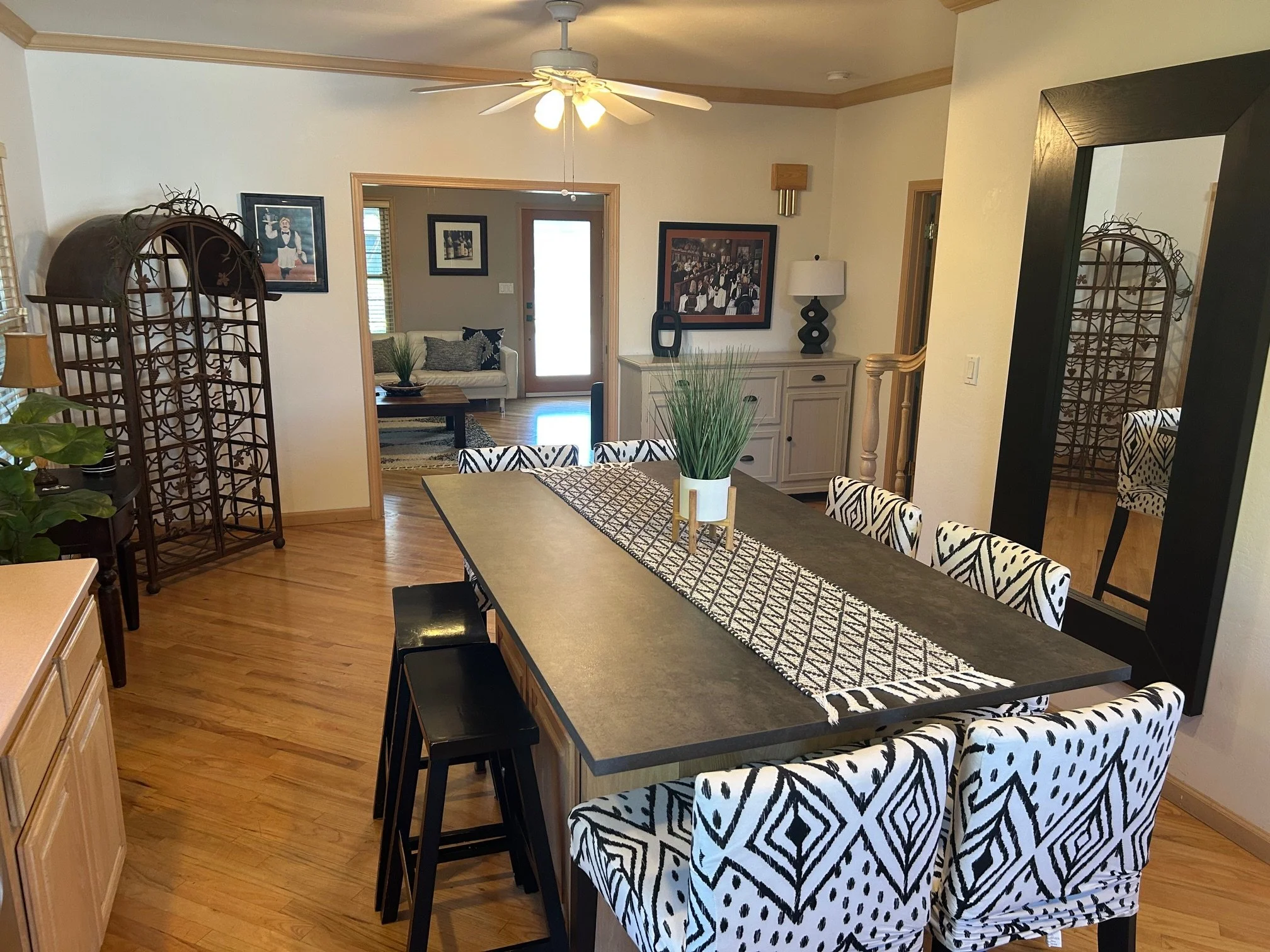 A dining room with a long black table, black and white patterned chairs, a decorative plant in a white vase, hardwood flooring, a large mirror on the wall, a sideboard with decor, a ceiling fan, and a view into a living room with a couch and door to the outdoors.