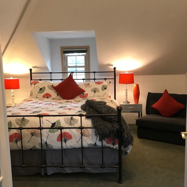 A cozy attic bedroom with a wrought iron bed, floral bedding, red accent pillows, and matching red bedside lamps. There's a window in the sloped ceiling, and a gray armchair with a red pillow beside the bed.