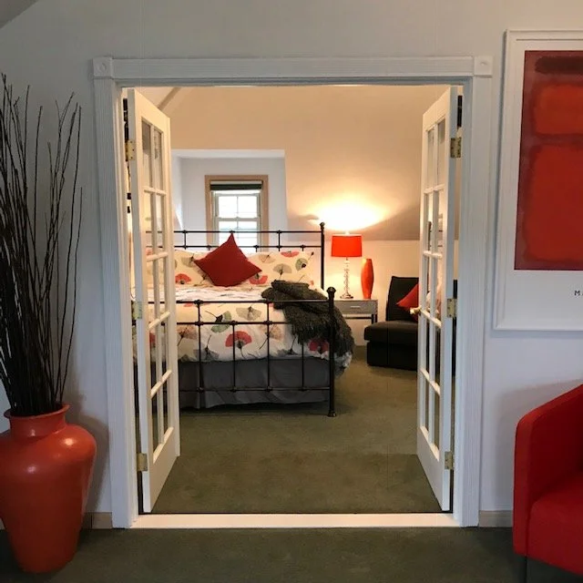 View through open French doors into a cozy bedroom with a metal bed frame, floral bedding, red pillows, a nightstand with a red lamp, and a black armchair. There's a window on the back wall, green carpet, and a painting on the right wall.