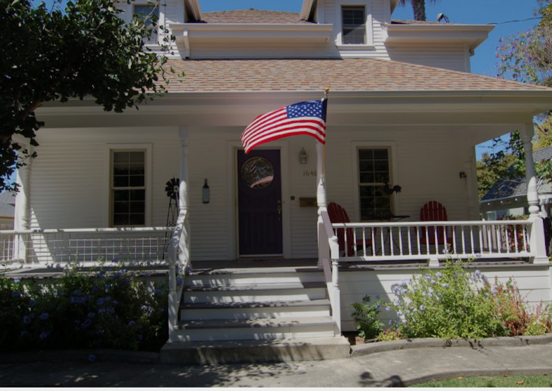 The Wine Maker's Estates is a charming, white house with purple door, American flag, porch chairs, and well appointed front yard.