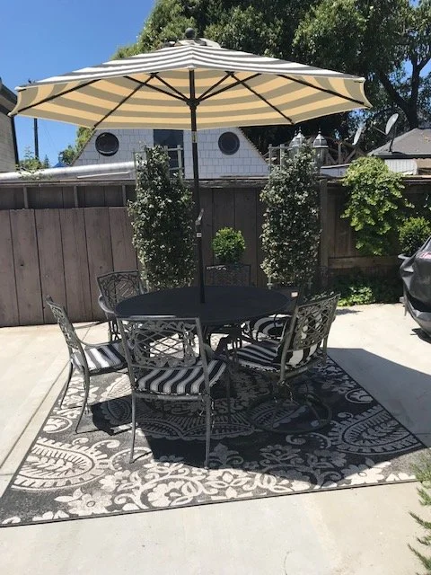 Patio setup with a round black table, six metal chairs with striped cushions, a large beige and black striped umbrella, a decorative outdoor rug, and potted plants against a wooden fence.