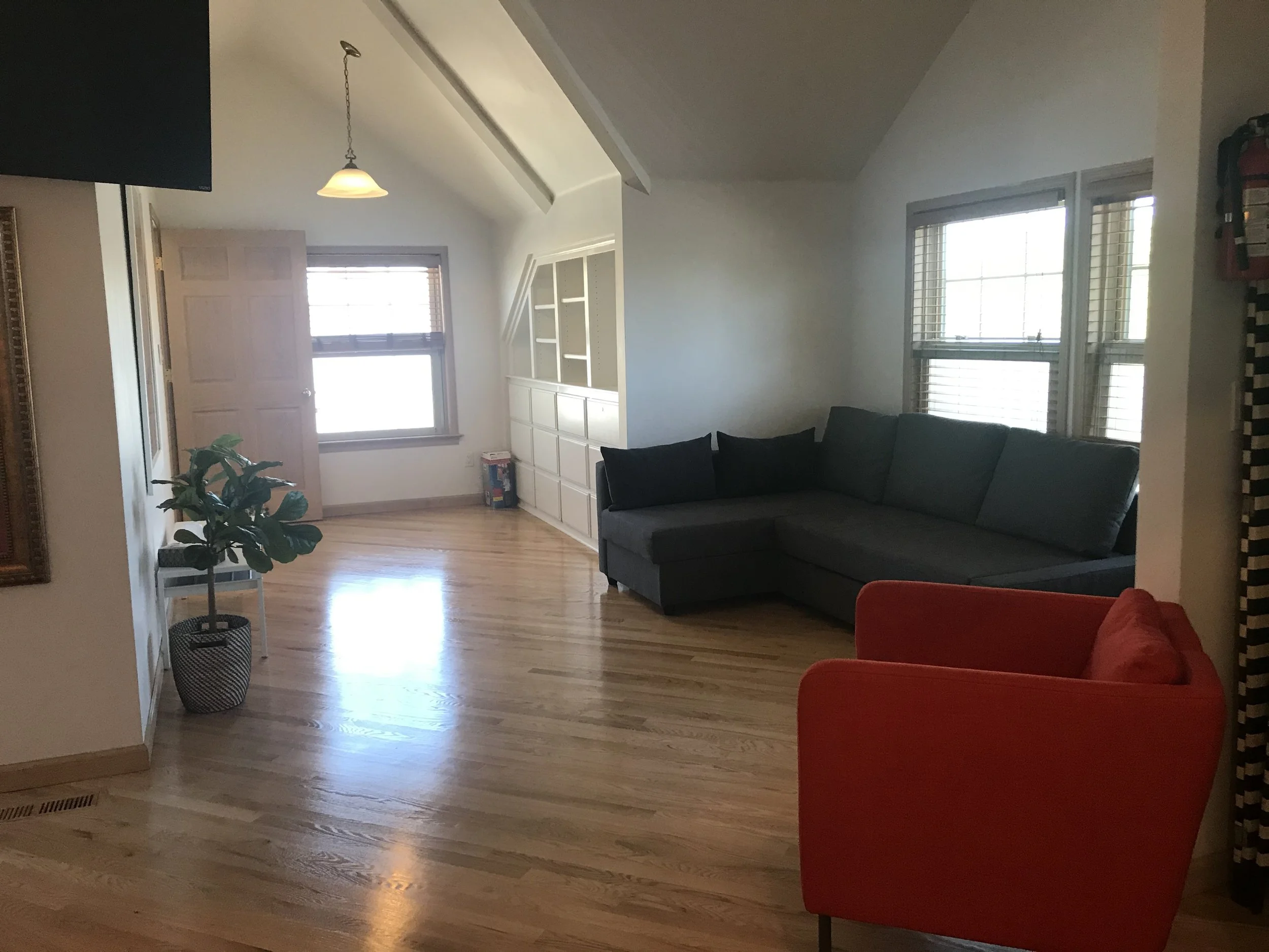 Living room with hardwood floor, gray sectional sofa, red armchair, potted plant, built-in white shelving, three windows with blinds, ceiling with sloped roof, and a hanging light fixture.