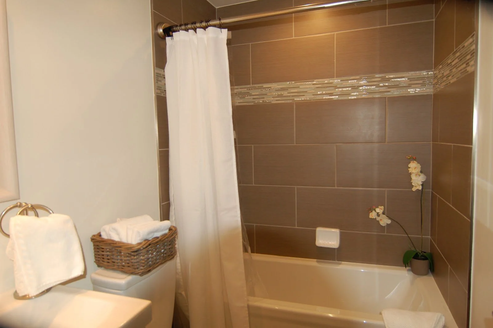 Bathroom with a shower and bathtub, white shower curtain, brown tiled wall with decorative trim, orchid plant, and towel.