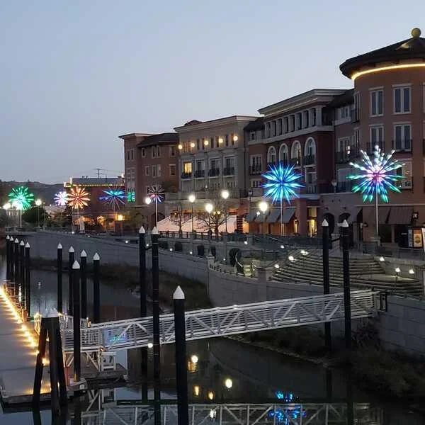 Nighttime scene along a canal with colorful firework-shaped lights on buildings and street lamps illuminating a walkway with stairs.