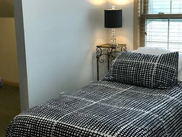 A bedroom with a bed covered in black and white geometric patterned bedding and a matching pillow. There is a small decorative table with a black lamp next to the bed and a window with blinds.