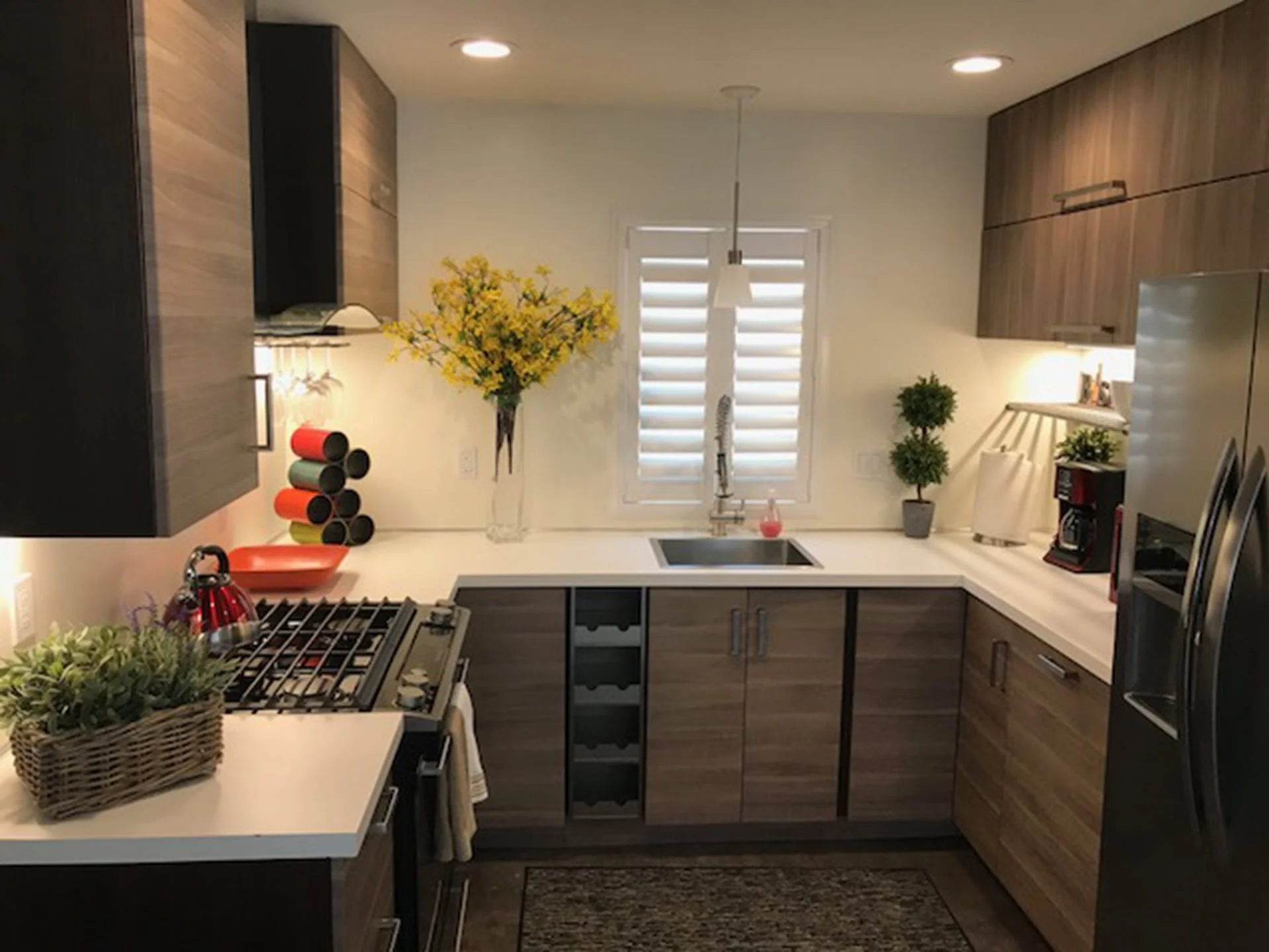 Modern kitchen with wooden cabinets, white countertops, a stainless steel sink, and decorative plants. There is a window with shutters, a coffee maker, and colorful mugs. A basket of green plants and a vase with yellow flowers are also visible.