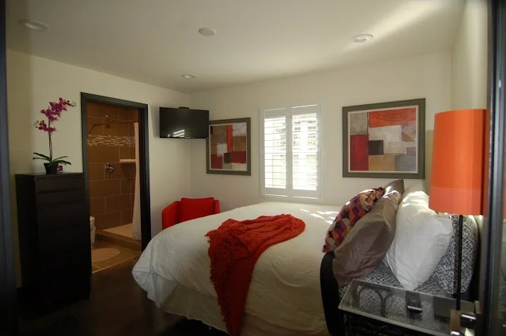 A bedroom with a bed covered in white bedding, a red throw blanket, and colorful pillows. The room has two framed abstract paintings, a window with shutters, a flat-screen TV, and a bathroom with a shower visible through an open door. There's also a tall lamp, a black dresser with a potted purple orchid, and a small red armchair.