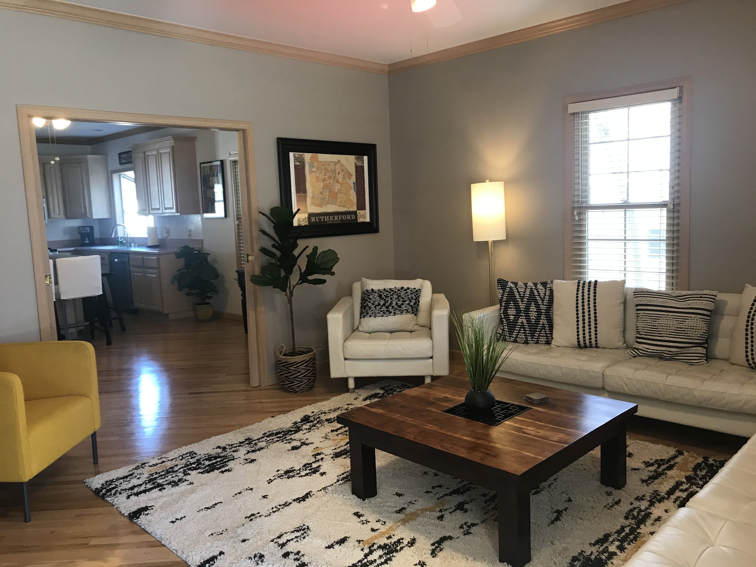 Living room with a white sofa, a white armchair, a yellow armchair, a wooden coffee table with a potted plant, a framed map of Rutherford, and a window with blinds. The room has a light blue wall, a tall floor lamp, and a black and white patterned rug.