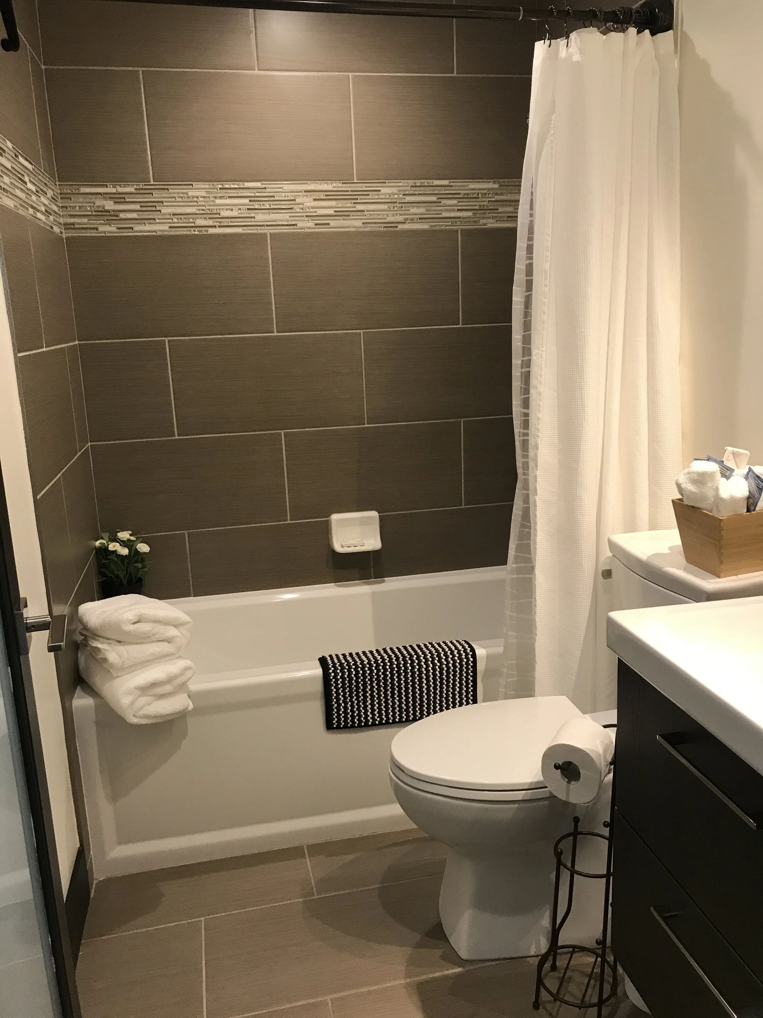 A bathroom featuring a bathtub with a white towel, a potted plant, and a black and white striped towel hanging on the tub's edge. The wall is tiled with large gray tiles and a decorative horizontal stripe. To the right is a toilet with a roll of toilet paper, a sink with a dark cabinet, and a wooden box with rolled towels on top. A white shower curtain and various toiletries are also visible.
