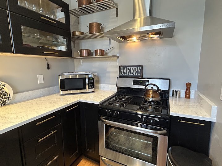 Modern kitchen with black cabinets, stainless steel oven and microwave, open shelving with copper pots, a stainless steel range hood, a bakery sign, and a teapot on the stove.